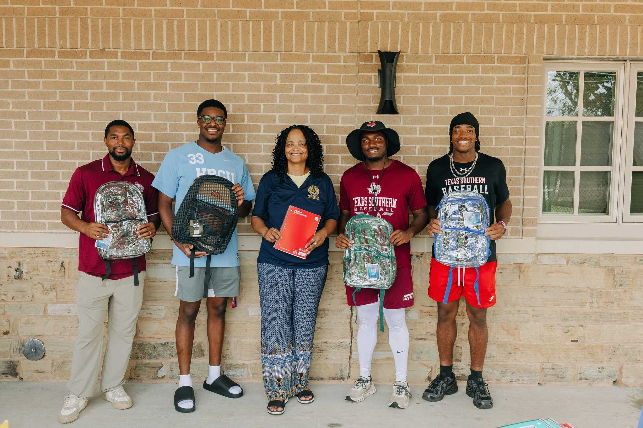 Five people stand outdoors holding backpacks and school supplies in front of a brick building, participating in a community back-to-school donation or distribution event.
