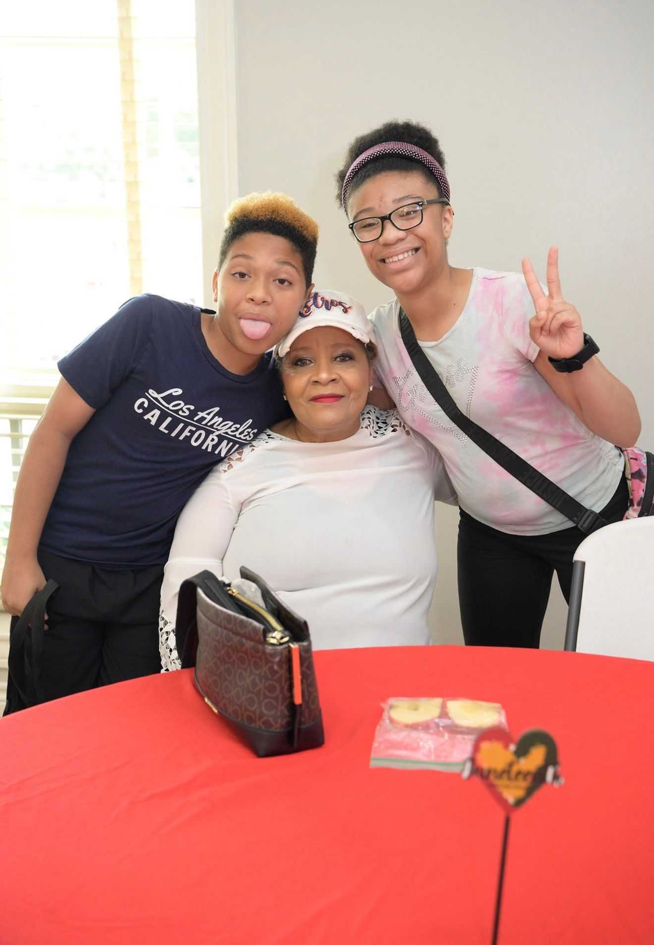 Adult seated at a table with two younger people leaning in; one holds up a peace sign while snacks and a purse rest on a red tablecloth.