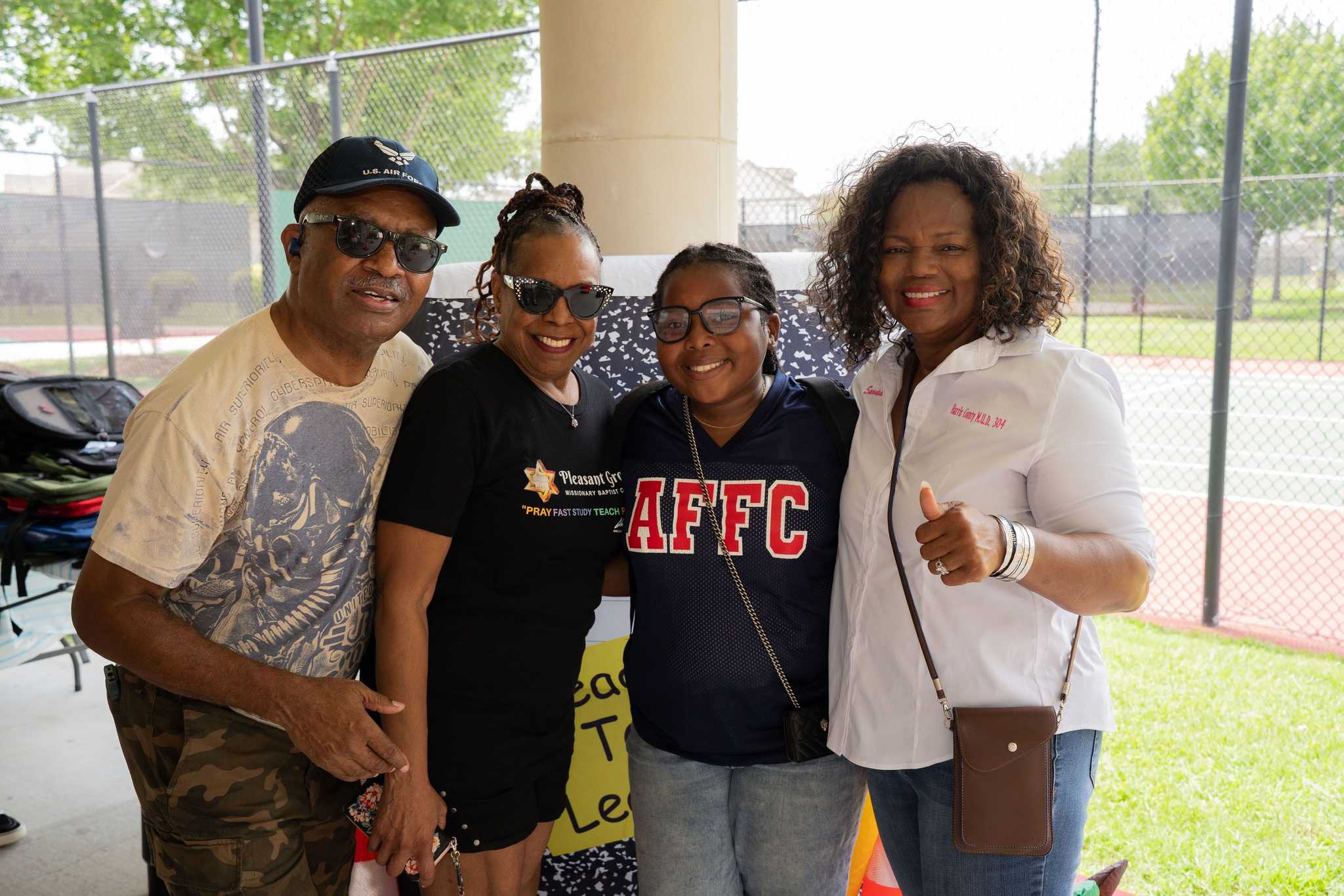 Four community members pose together at a back-to-school event near tennis courts, standing in front of school-themed decorations and backpack distribution tables.