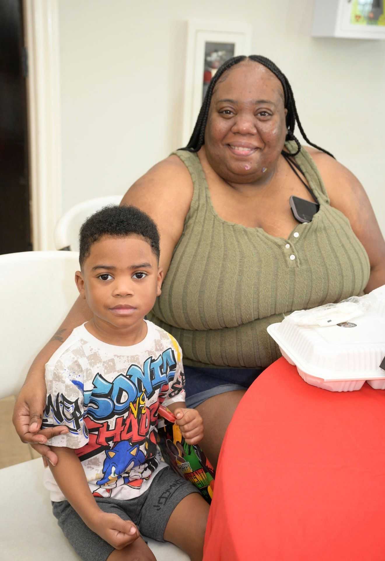 Adult and young child sit at a table with takeout container and bag, posing together indoors at a community event.