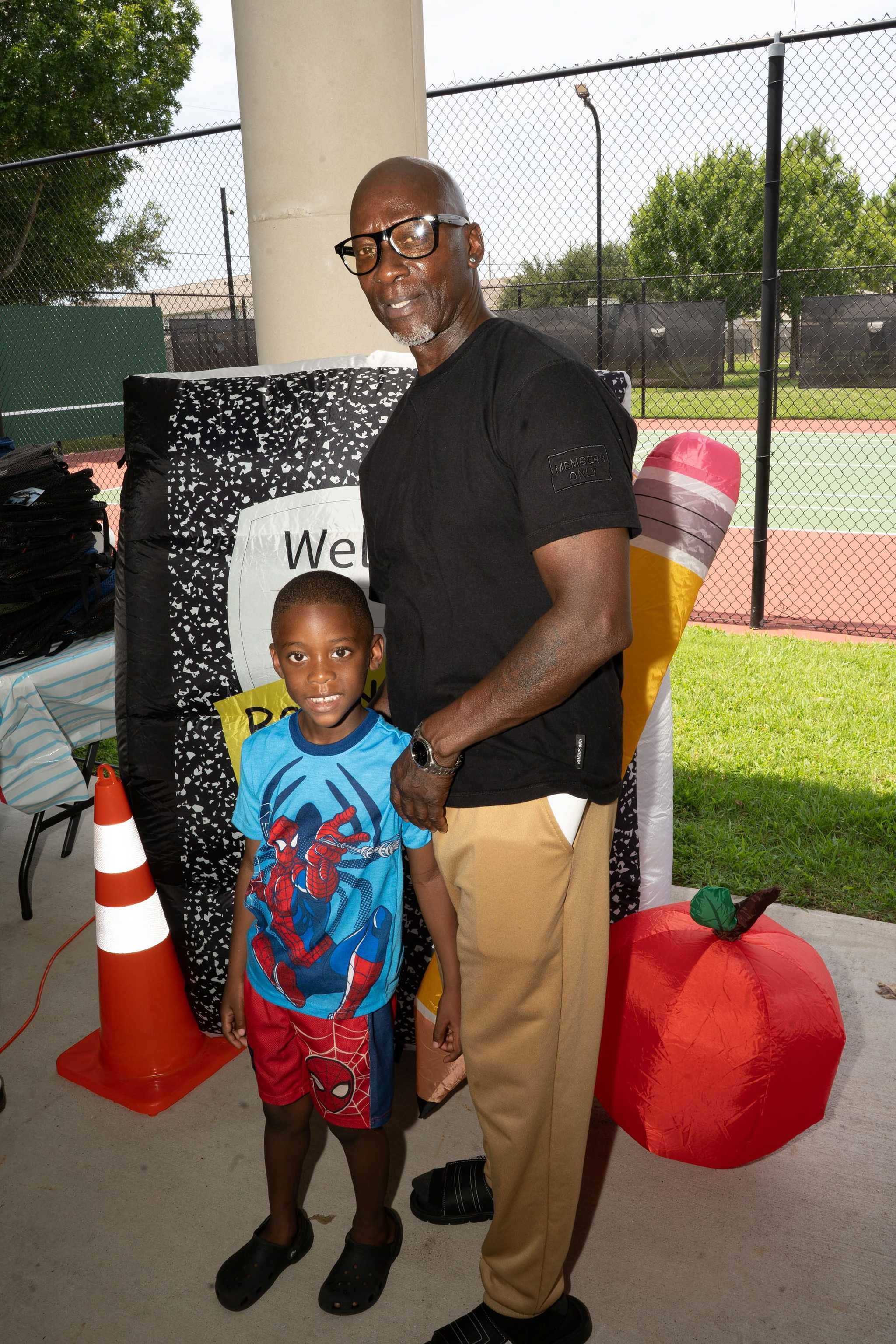 Person and child pose at a community event beside school-themed decorations, including an oversized pencil, notebook display, traffic cone, and apple near tennis courts.