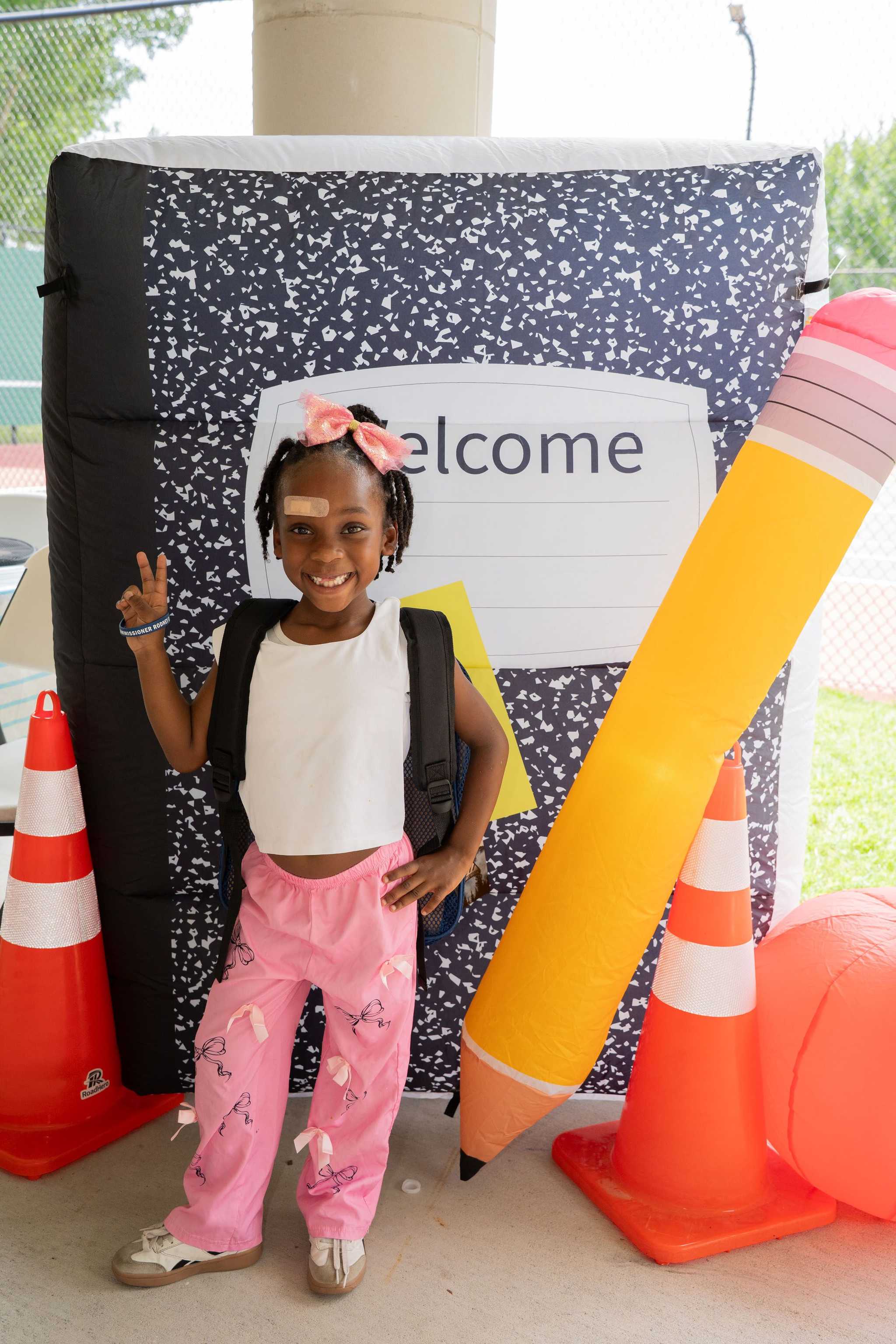 Four community members pose together at a back-to-school event near tennis courts, standing in front of school-themed decorations and backpack distribution tables.