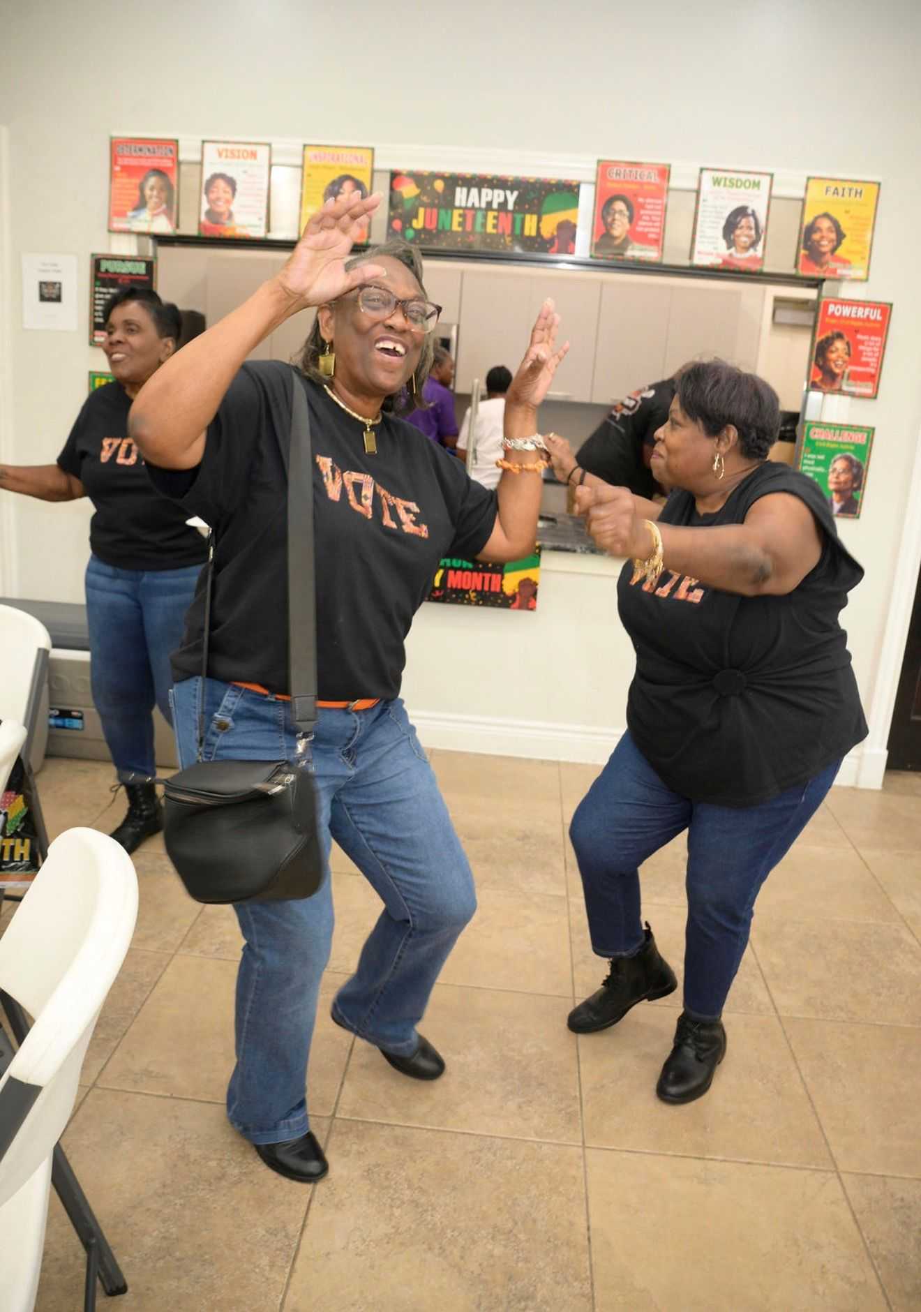 Two people dance playfully indoors at a Juneteenth event, wearing matching shirts with posters and a “Happy Juneteenth” banner in the background.