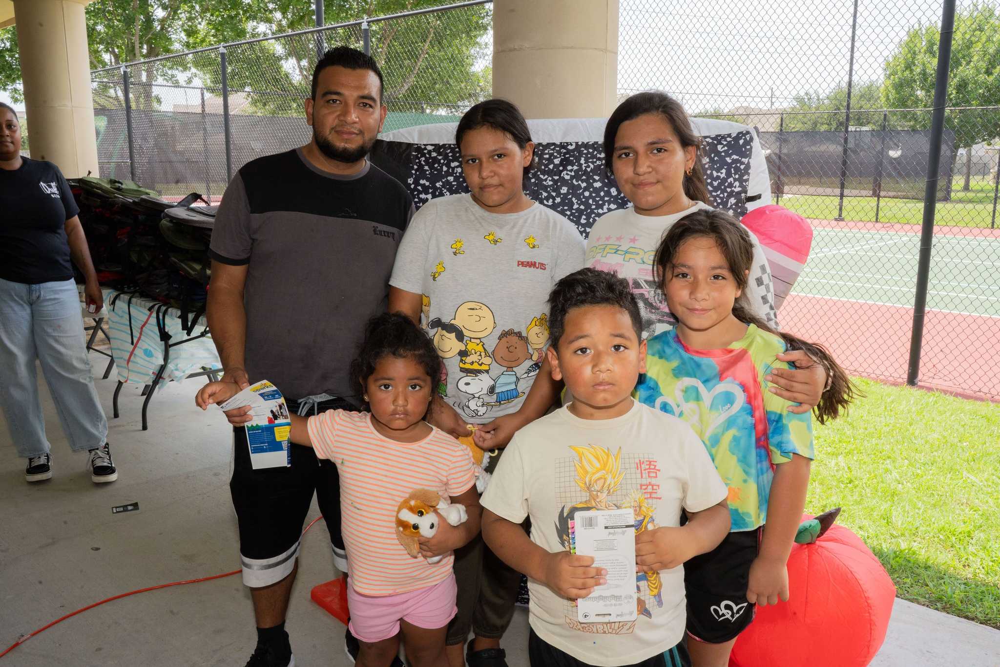 Group of adults and children pose at a community back-to-school event near tennis courts, holding informational handouts beside oversized school-themed decorations.