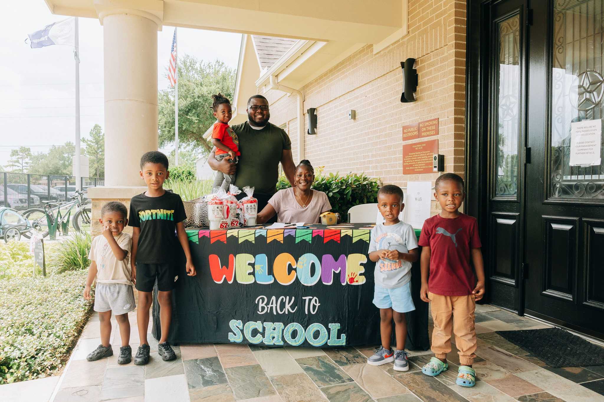 Four community members pose together at a back-to-school event near tennis courts, standing in front of school-themed decorations and backpack distribution tables.