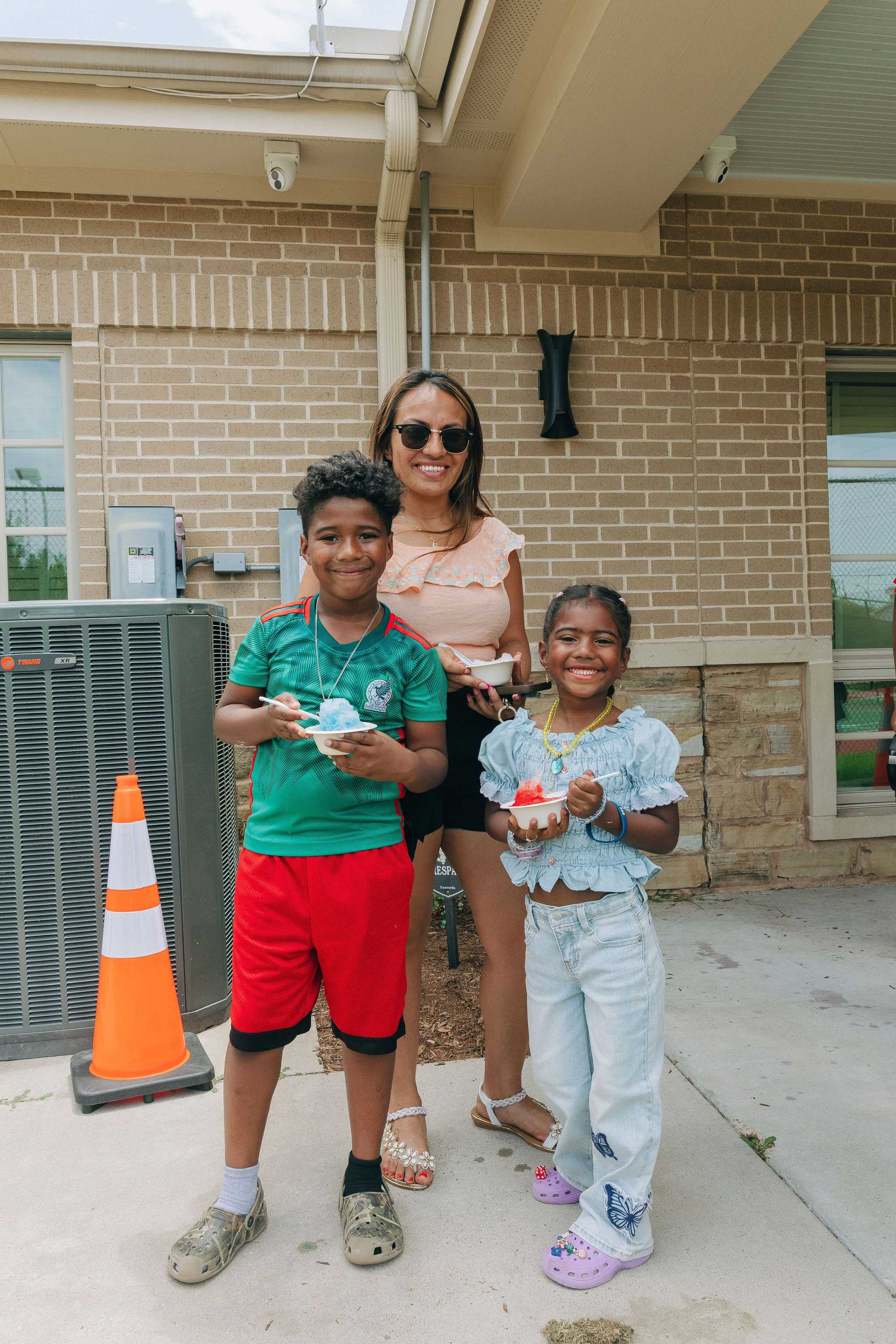 Three people stand outdoors holding cups of shaved ice near a community building, enjoying refreshments during a back-to-school event.
