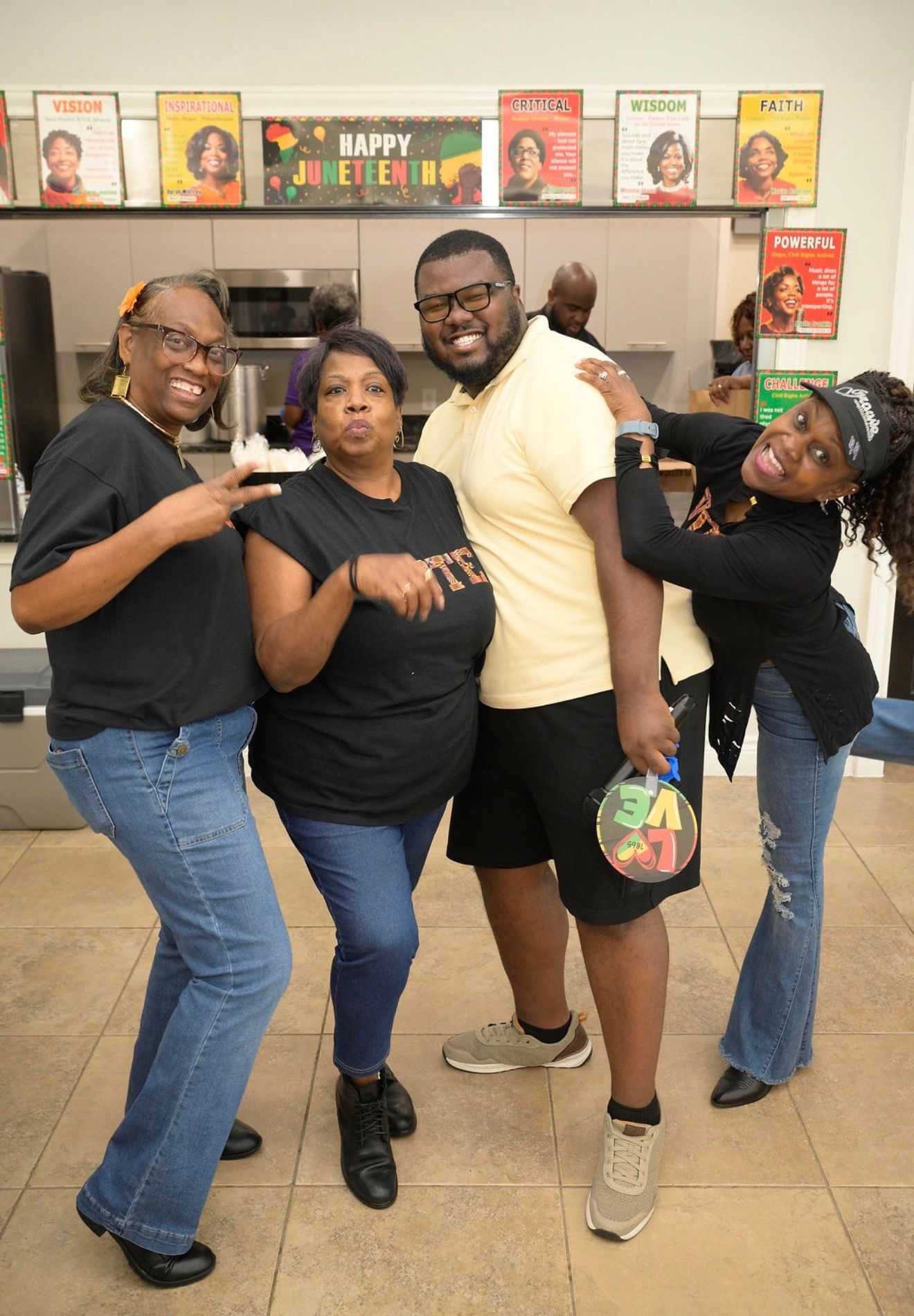 Four adults pose together indoors in a kitchen, smiling and standing in front of a “Happy Juneteenth” banner and colorful posters.