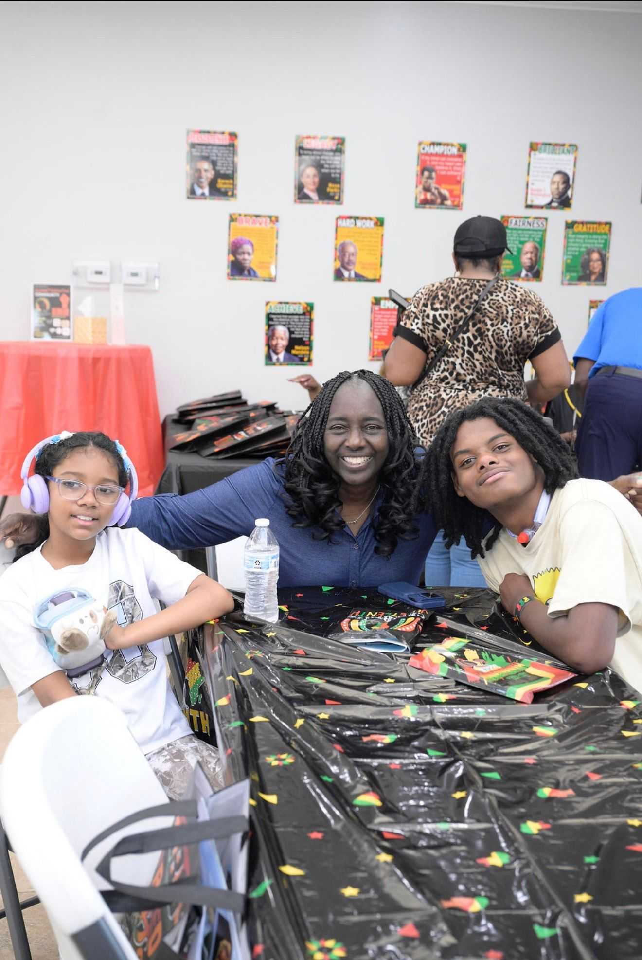 Three people sit together at a table with Juneteenth decorations, including a patterned tablecloth and snack items, with posters displayed on the wall behind them.