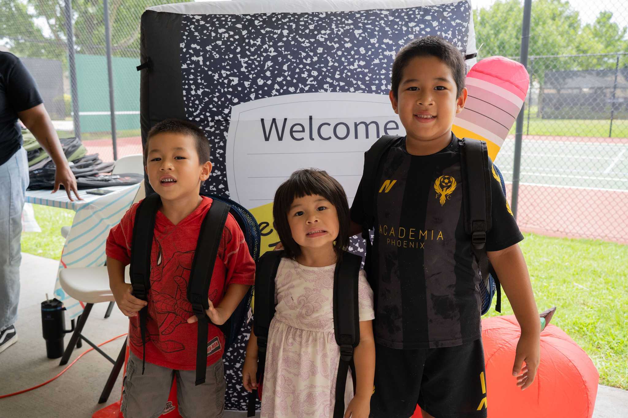 Three children wearing backpacks pose in front of oversized school-themed decorations, including a notebook, pencil, and apple, at a community back-to-school event.