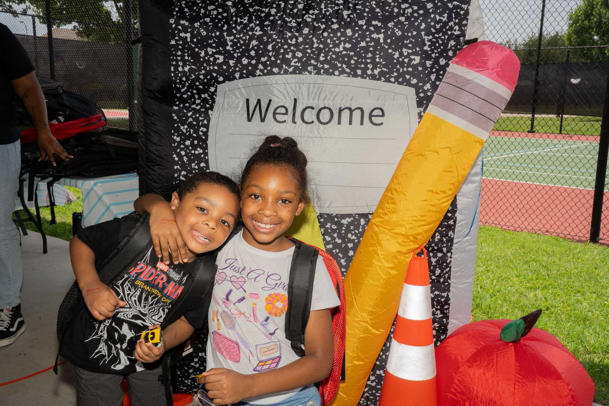 Two children wearing backpacks pose beside oversized school-themed decorations, including a notebook, pencil, traffic cone, and apple, at a community back-to-school event.
