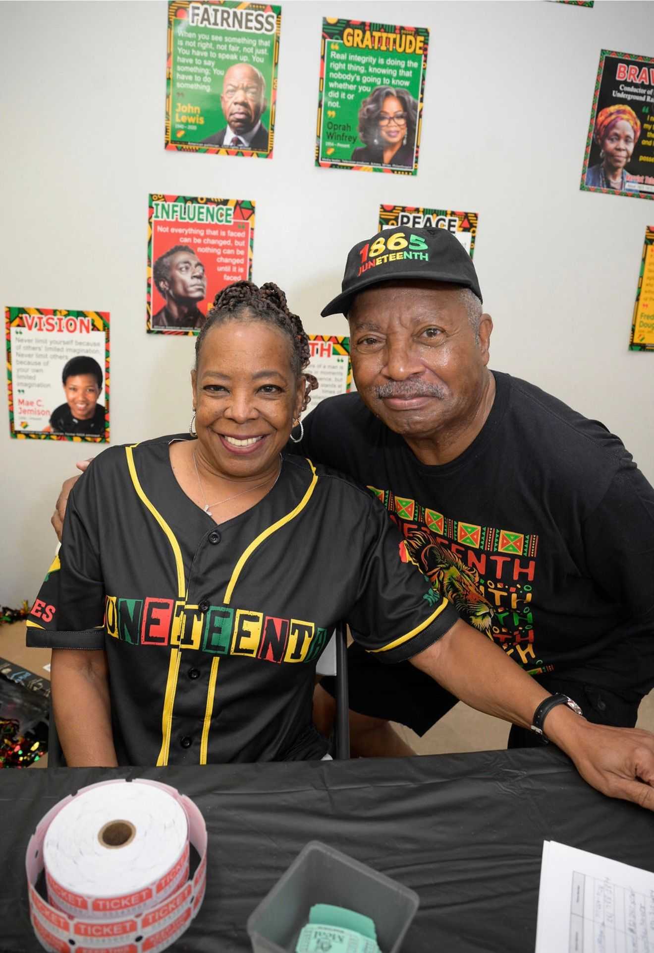 Two adults wearing Juneteenth apparel sit at a table with raffle tickets, posing in front of a wall decorated with inspirational posters.