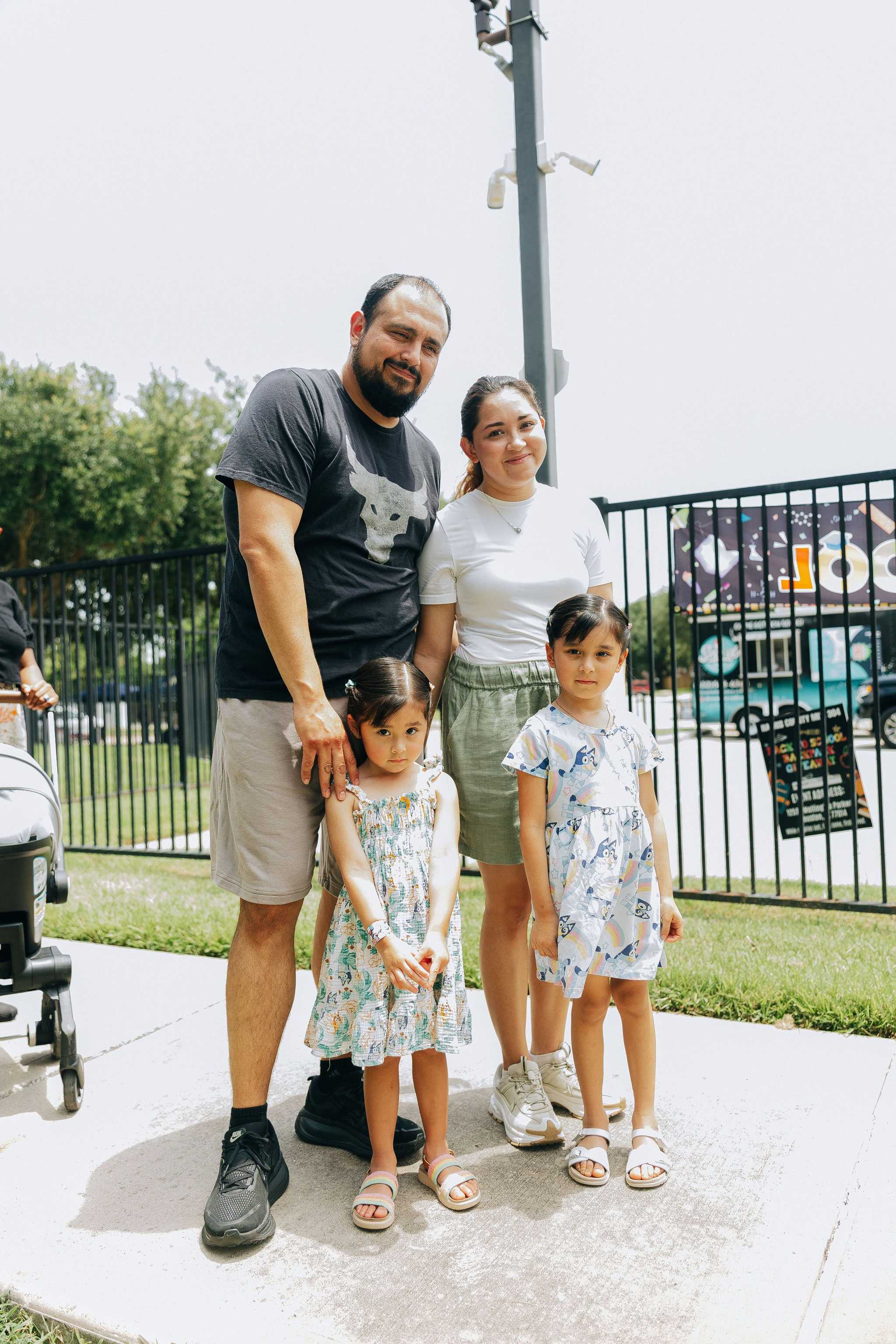 Four people stand together on a sidewalk near a fenced recreational area, with security cameras mounted on a pole in the background and outdoor amenities visible nearby.